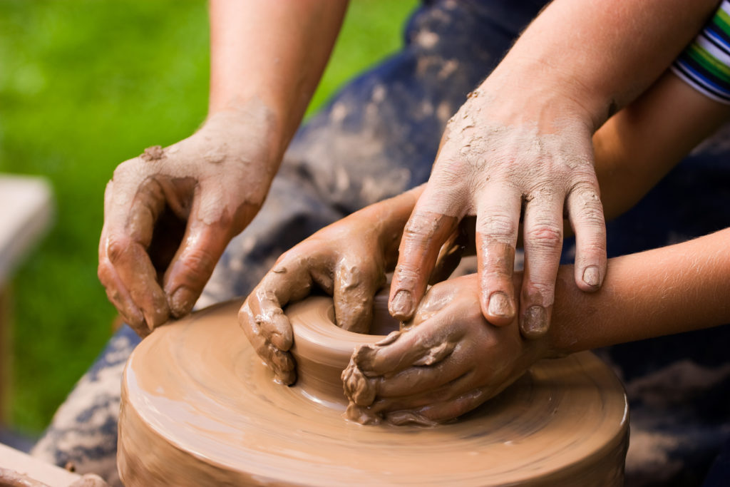 A potters hands guiding a child hands to help him to work with the ceramic wheel